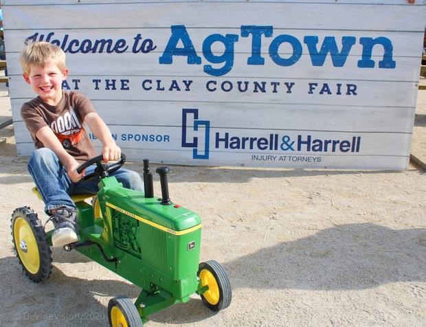Young boy rides green toy tractor at AgTown sign.