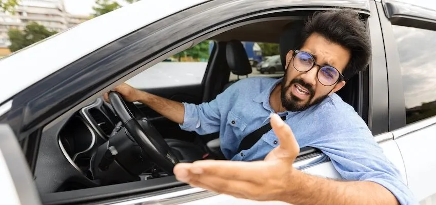 Man with glasses in blue shirt leaning out car window arguing.