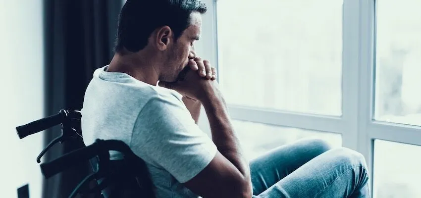 Man in wheelchair sits by window, elbows on knees.