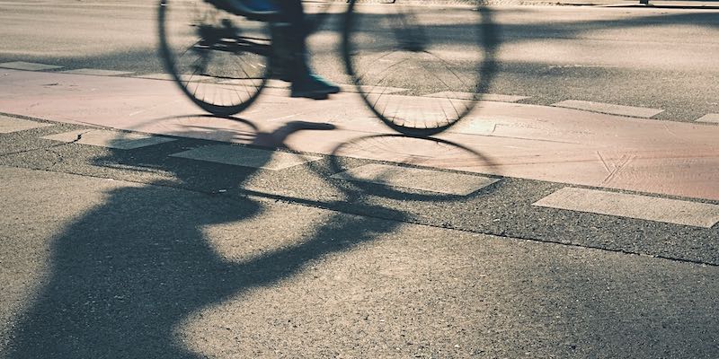 Cyclist rides on sunlit street casting shadow.