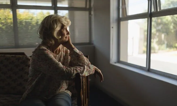 Older woman rests chin on hand, gazes out window.