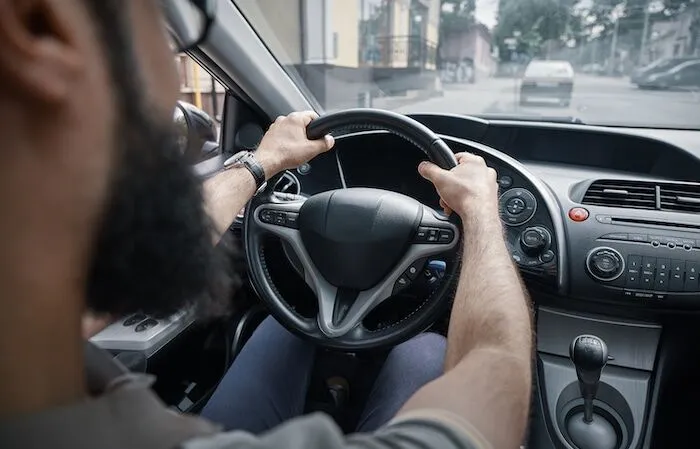 Bearded person driving car with both hands on wheel.