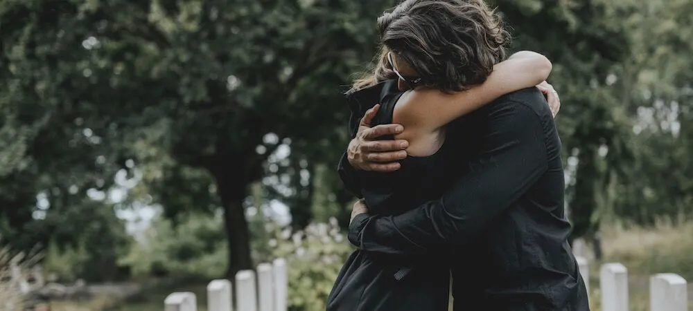 Two people in black embrace in a cemetery.