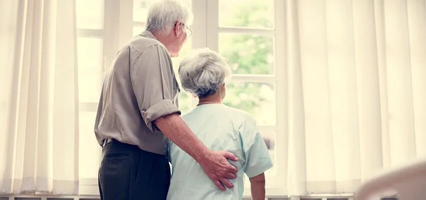 Elderly couple stands by window looking outside.