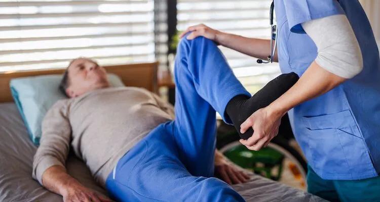 Healthcare worker lifts and bends older adult’s leg in bed.