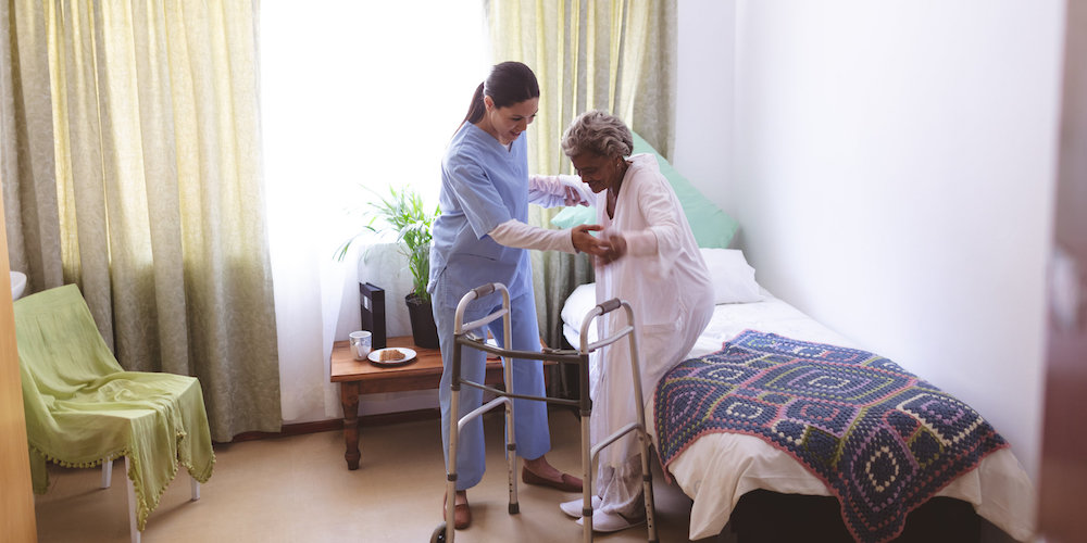 A nurse helps an elderly person stand using a walker.