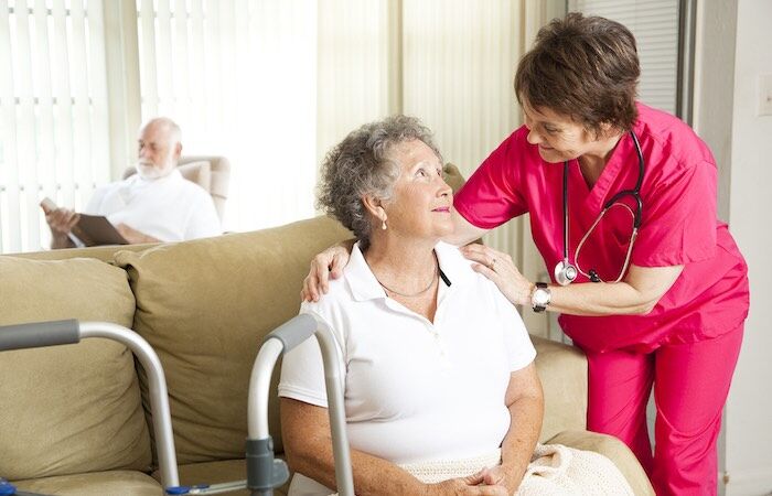 Nurse chats with elderly woman as man reads nearby.