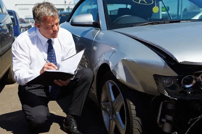 Man in shirt and tie kneels by damaged silver car.