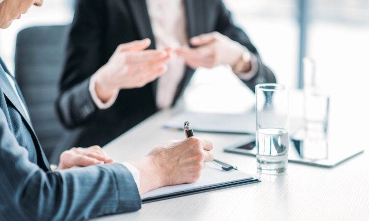Two people at a desk, one writing, one gesturing.
