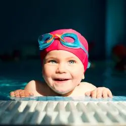 Smiling baby in red swim cap and blue goggles rests.
