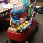 A red wagon filled with boxed toys sits on carpet.