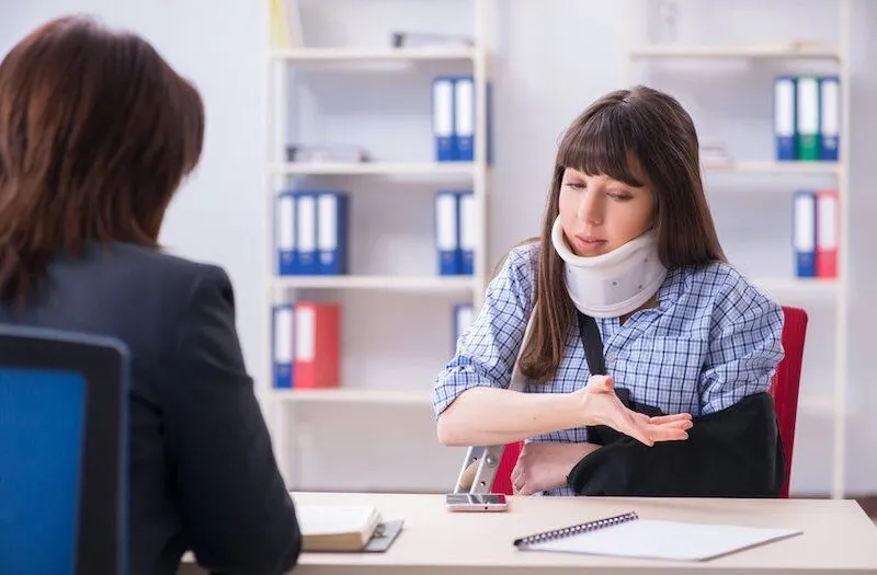 Woman in neck brace and arm sling talks at desk.