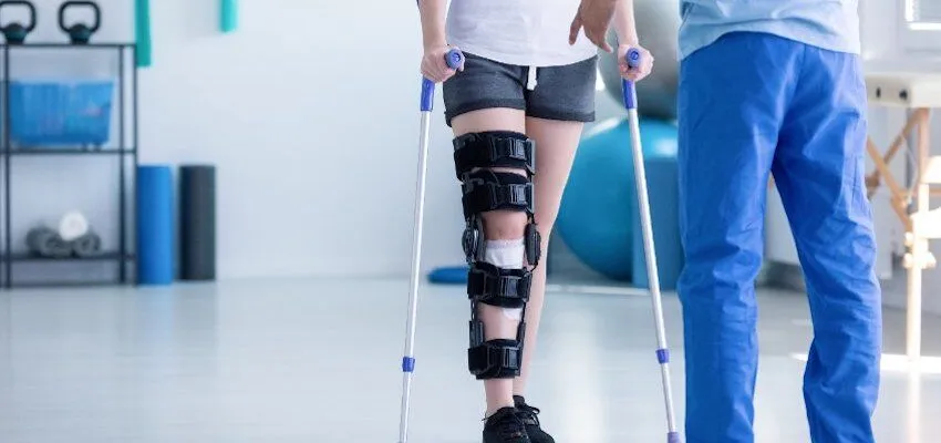 Person with a knee brace uses crutches alongside healthcare professional.